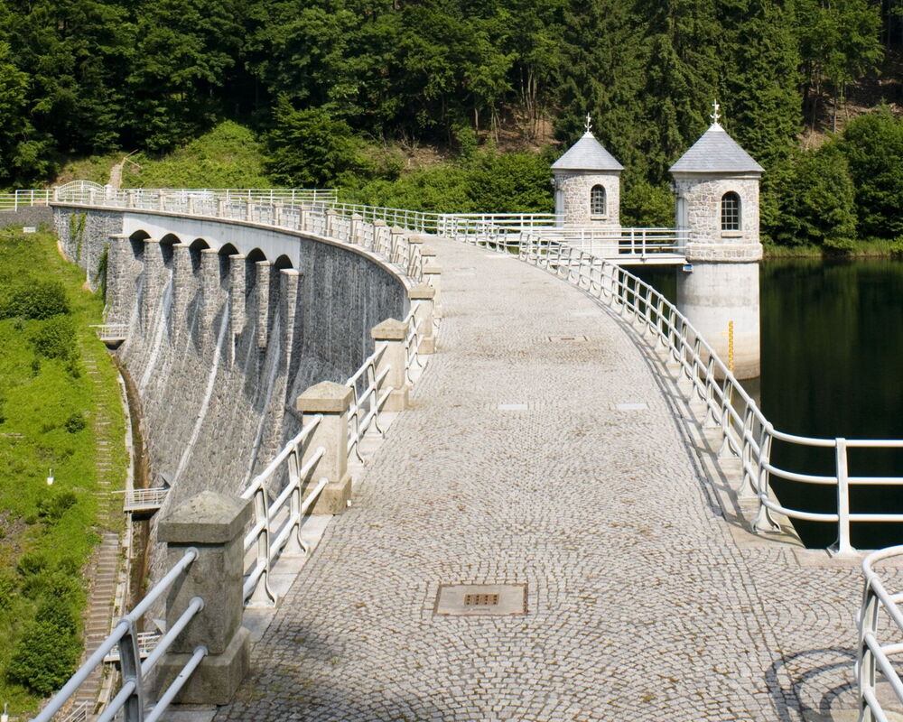 Seitenansicht auf das Absperrbauwerk der Trinkwassertalsperre Neustadt, die Mauerkrone ist gepflastert, mit einem Geländer umrandet und zwei Wasserentnahmetürme sind über Laufstege angebunden.