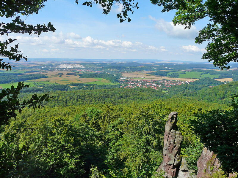 Blick auf eine Felsformation, im Hintergrund Laubwald, ein weißer Gipstagebau und eine Ortschaft im Sommer