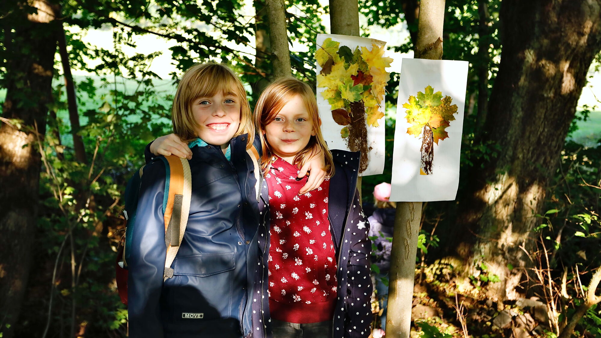 Zwei lächelnde Kinder stehen im Wald, neben ihnen hängen zwei selbstgemachte Blättercollagen